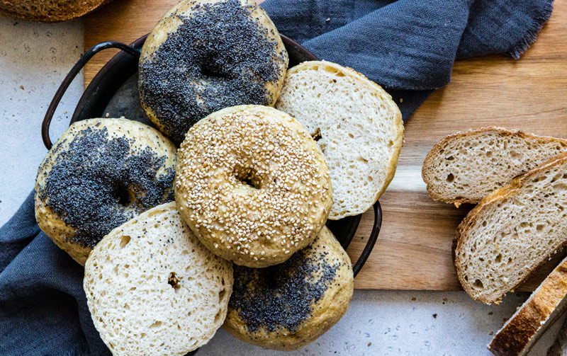 Des bagels sans gluten Piaf parsemés de graines de sésame et de pavot dans un plat sur un torchon et quelques tranches de pain sans gluten Piaf sur le côté